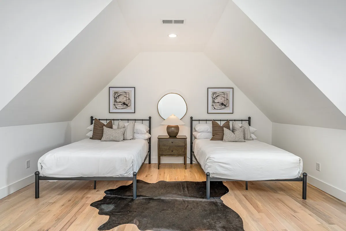 A bedroom featuring two full beds, a stylish round mirror above a nightstand with a lamp, and vibrant artwork on the walls. The scene is relaxed and inviting, suggesting a perfect spot for siblings or friends to unwind together.
