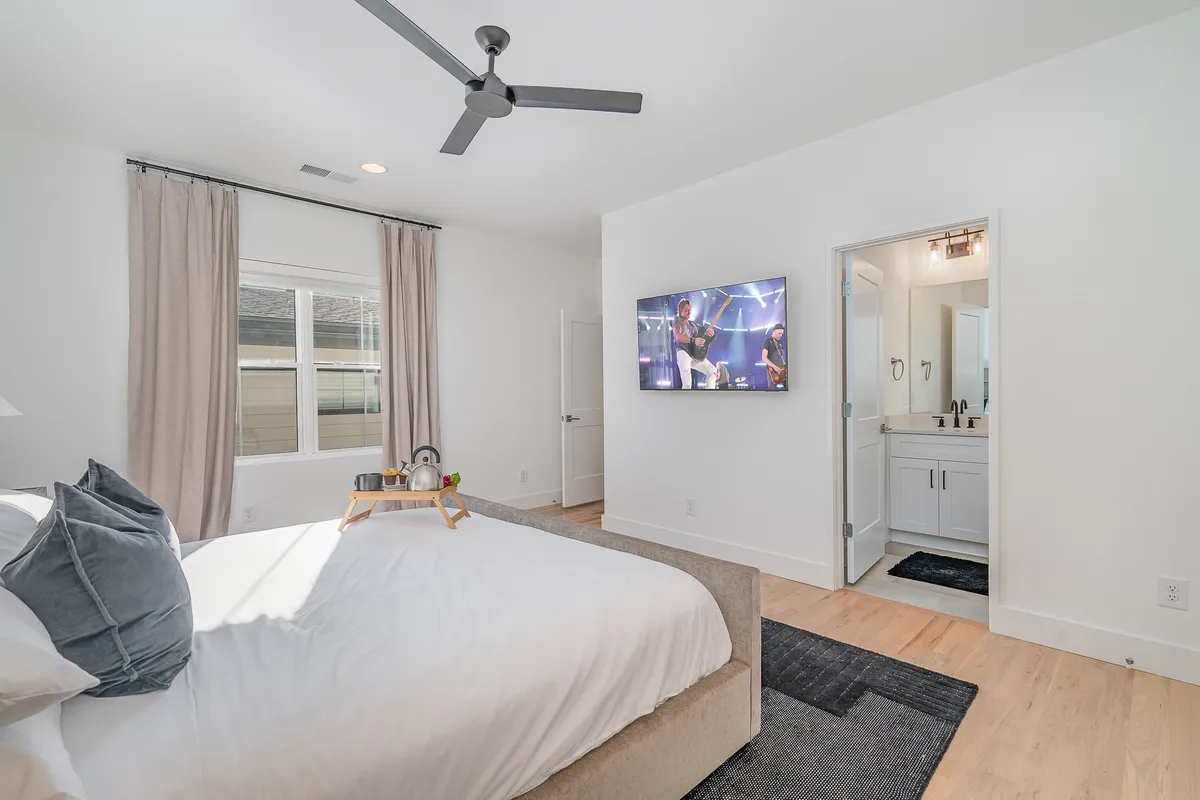 A modern bedroom with a king-sized bed adorned with decorative pillows, a smart TV mounted on the wall, curtains framing a sunlit window, and a bathroom door visible to the side. A tray with morning drinks sits on the bed, and soft lighting creates a cozy, comforting atmosphere.