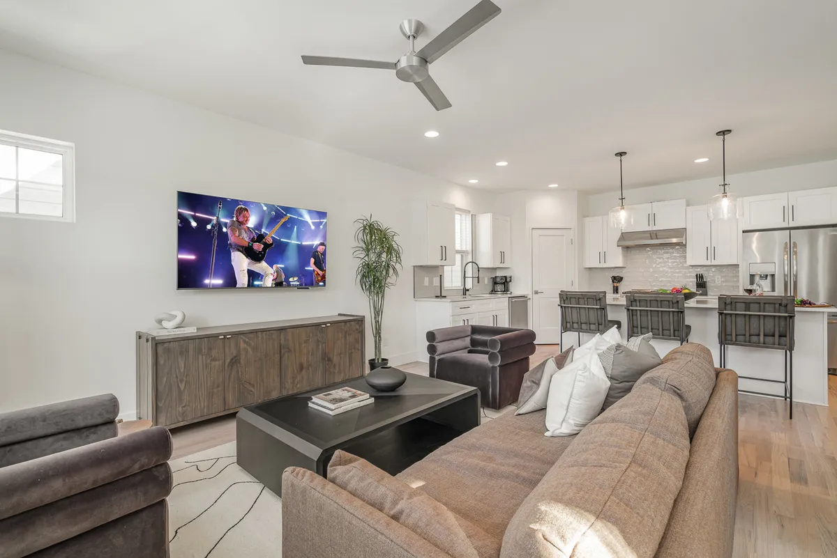 The image showcases a spacious, inviting living area featuring a large TV displaying a live concert, a plush and comfortable sofa perfect for lounging, and a stylish kitchen island set up for casual dining or entertaining. The layout is open and social, designed to bring together guests for relaxing movie nights, memorable concert viewings, or friendly gatherings.