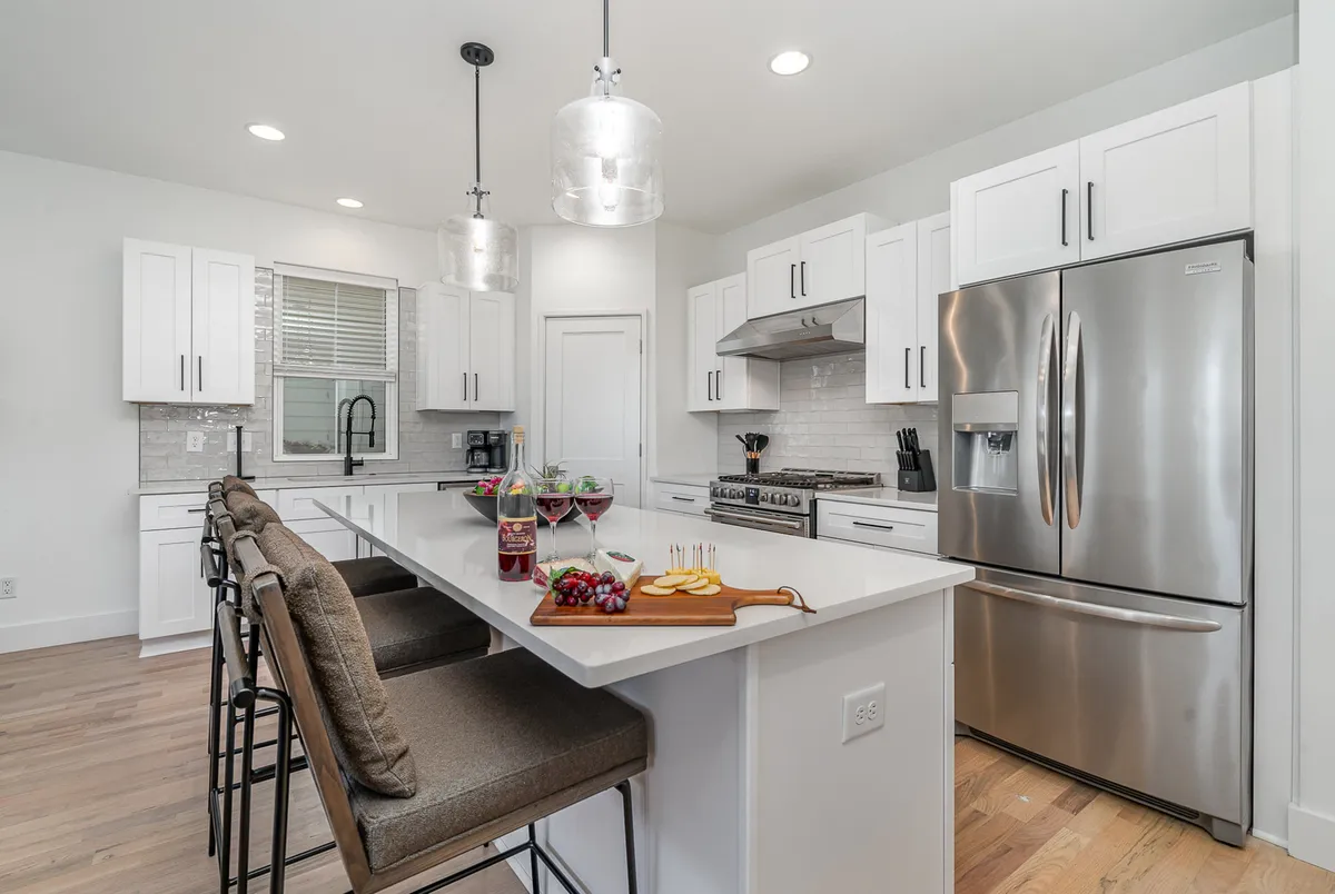 A modern kitchen with a central island countertop, bar stools, stainless steel appliances, and hanging light fixtures. A wooden cutting board with fresh food hints at meal preparation and social cooking, while warm tones and thoughtful design invite gathering and celebration.