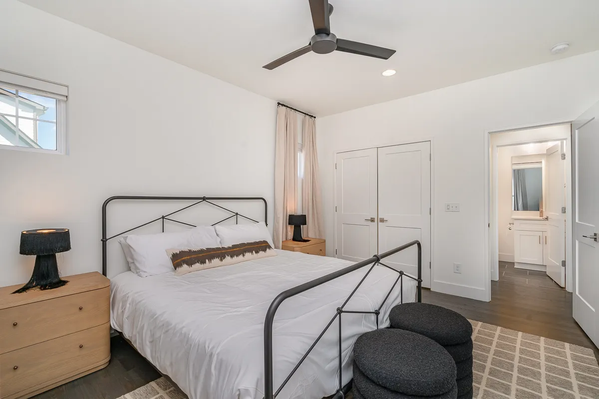 A serene bedroom with a large bed dressed in fresh linens, illuminated by natural light filtering through the window. Beside the bed is a reading nook with textured stools and a nightstand lamp, creating a peaceful, calming space.