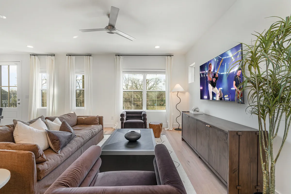 A spacious, contemporary living room featuring a large sectional sofa facing a wall-mounted flat-screen smart TV. The stylish coffee table, decorative plant, and modern light fixture create a comfortable and inviting atmosphere, enhanced by the natural light streaming in from nearby windows.