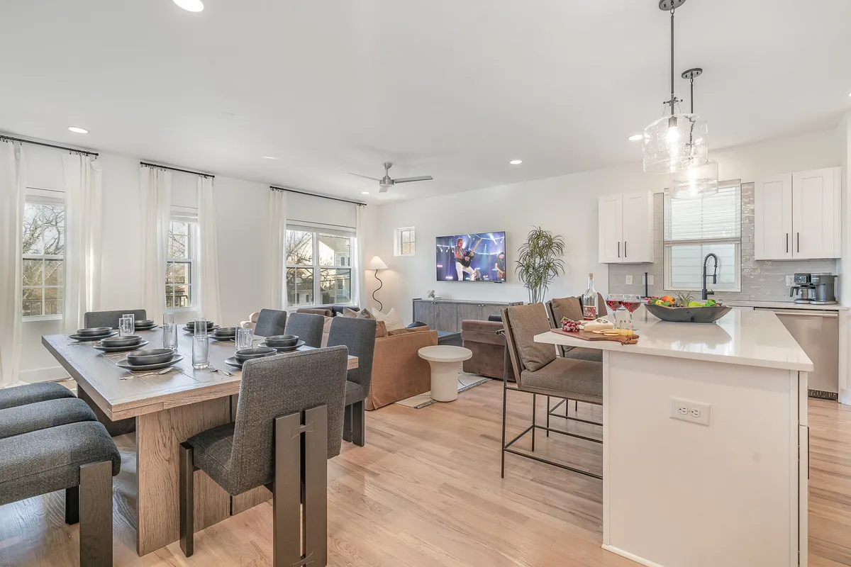 An open-plan space seamlessly connecting a comfortable dining table, sofa area, kitchen island, and visible television. The setting is warm, with inviting decor and ample seating, ideal for both shared meals and casual relaxation.